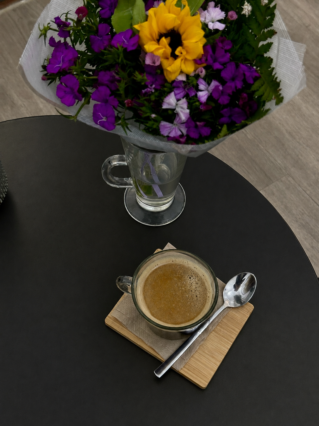 Taza de café sobre una base de madera y un pequeño ramo en un vaso, sobre una mesa redonda