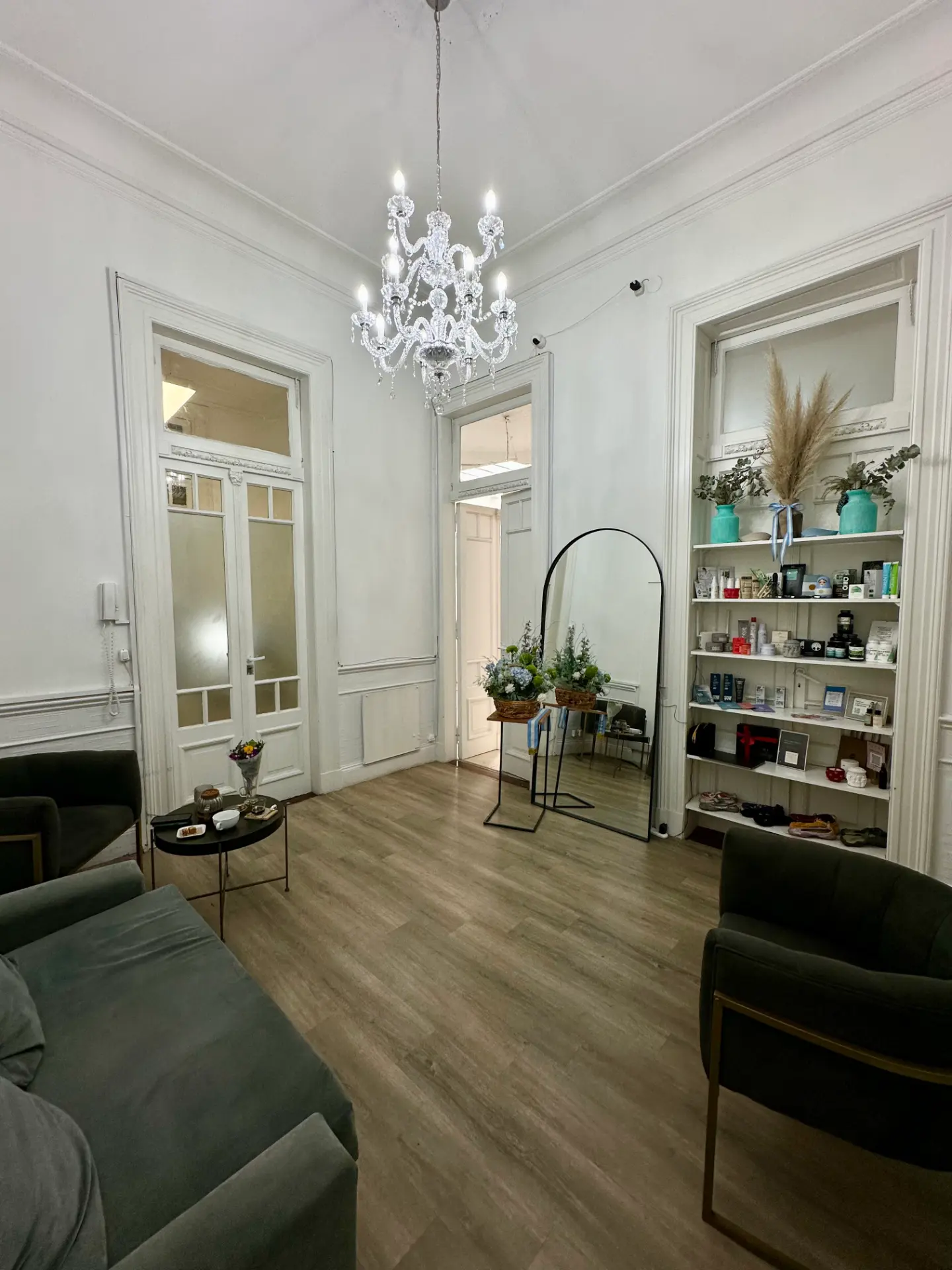 Sala de espera de La Mint: araña de cristal, techos altos con molduras, sofás y sillones grises, espejo con arco y estantería con cosmética