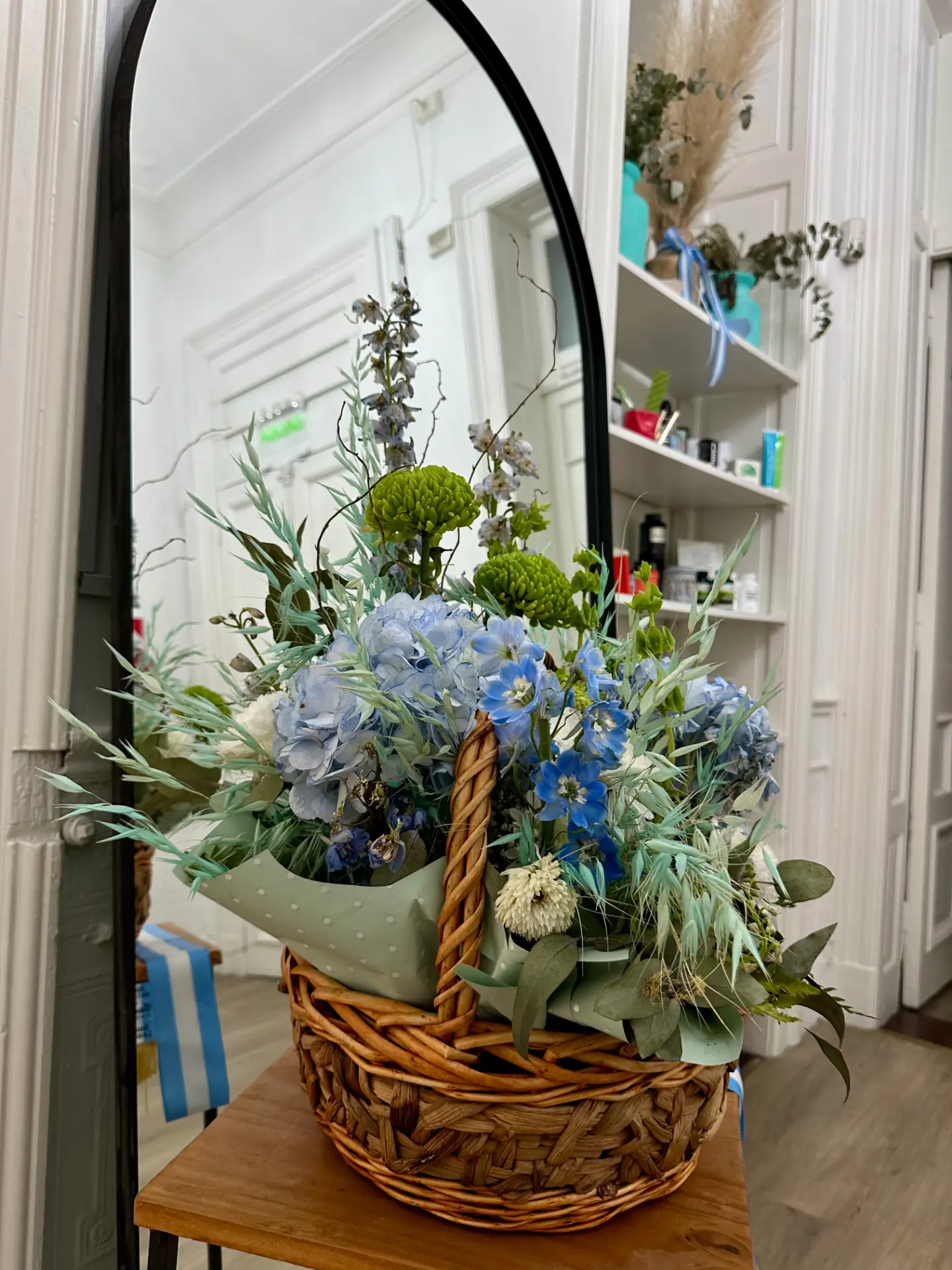 Canasta con hortensias azules, espuela de caballero y crisantemos verdes sobre una mesa de madera junto al espejo con arco